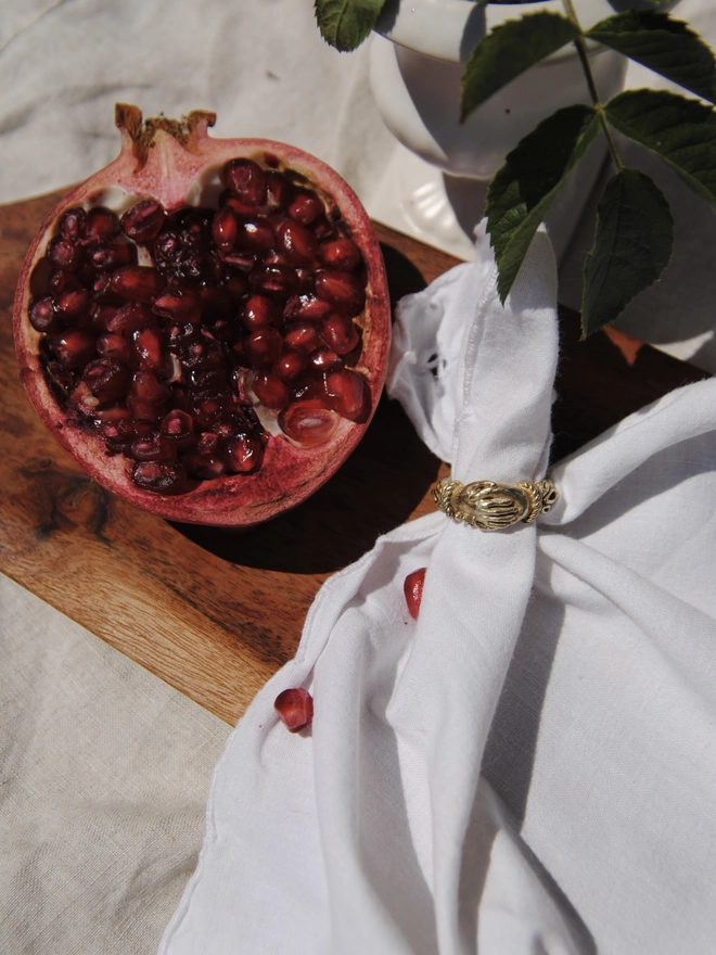 The image of a still life type layout including half a pomegranate, some leaves, a rustic piece of wood, and some white cloth, with a gold toned brass ring with 2 hands being held as the design laying on the white fabric 