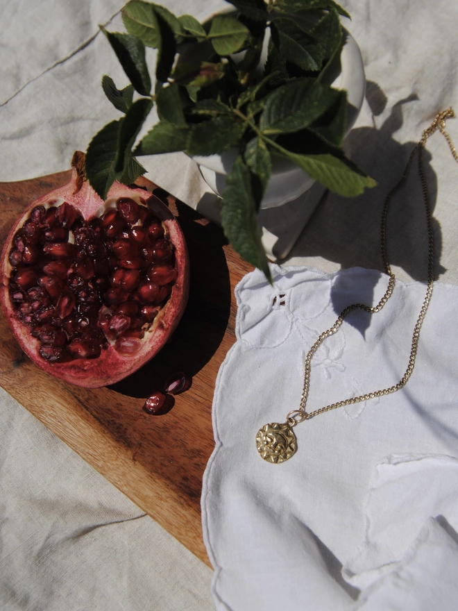 Image of a hand carved sun face pendant cast in gold toned recycled brass on a 9 carat gold chain. White vintage lace fabric under necklace, an a rustic wooden board, with half a pomegranate and rose leaves arranged in the image 