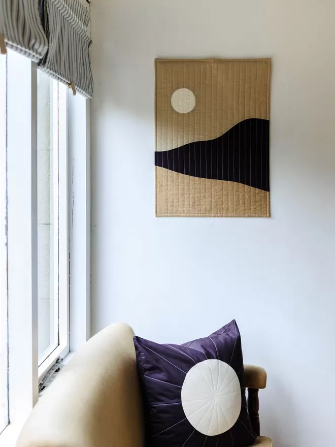 Moonrise Quilt Hanging On a White Wall Above a Chair and Next To a Window