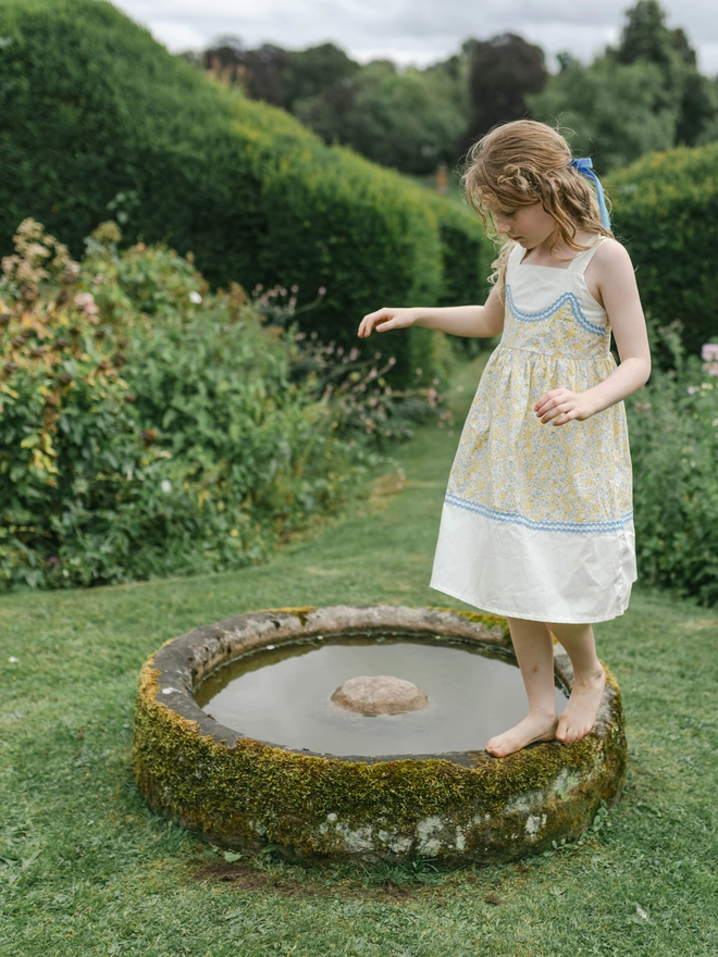A girl balances on an old trough wearing a yellow floral and yellow sundress with blue rikrak detail.