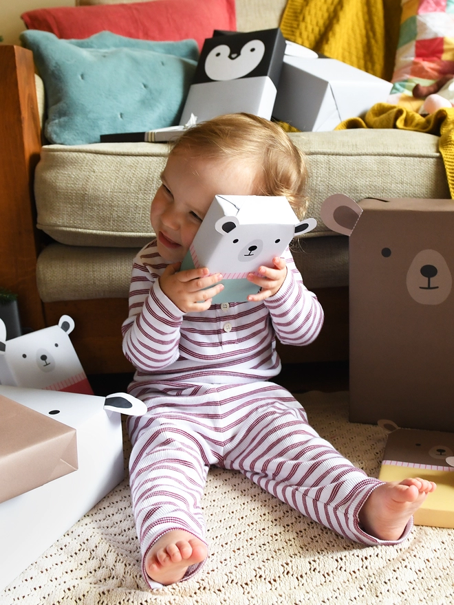 A baby wearing red and white striped pyjamas holds a gift wrapped as polar bear and is surrounded by other gifts wrapped in animal wrapping paper.