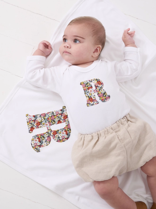 A baby laying on a soft white cotton blanket with an appliqué Liberty print initial on it