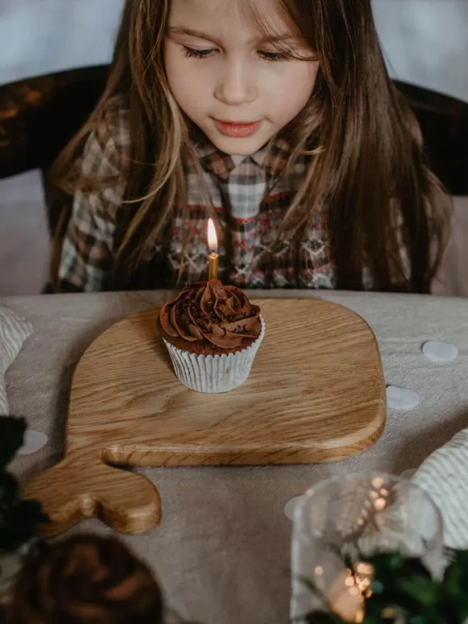 a chocolate cupcake with a lit candle resting on a whale wooden serving board with a little girl ready to blow the candle out 