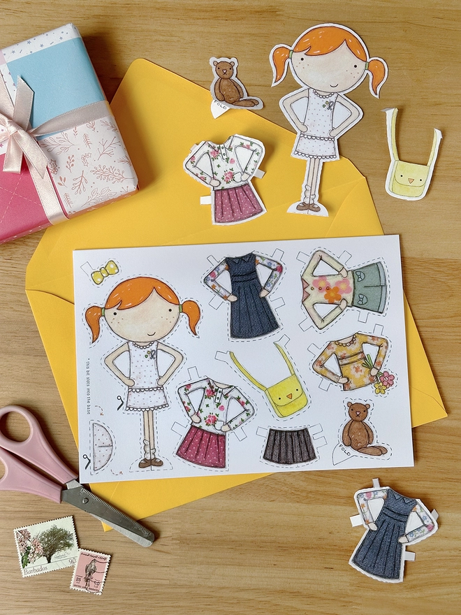 A paper doll greetings card with a paper doll and several outfits on the card lays on a yellow envelope on a wooden desk.