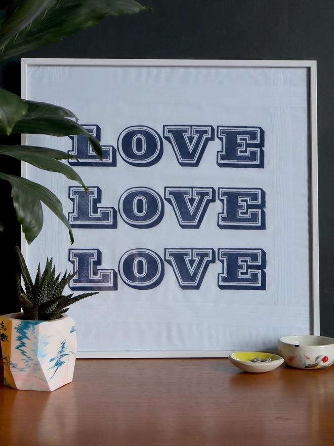 A Mr.PS Love hankie printed in midnight blue framed in a white picture frame propped on a sideboard in front of a dark wall