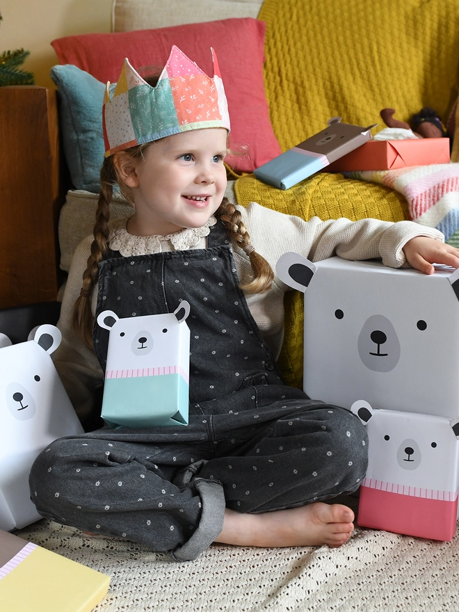 A young girl wearing grey dungarees and a patchwork crown holds a gift wrapped as polar bear and is surrounded by other gifts wrapped in animal wrapping paper.