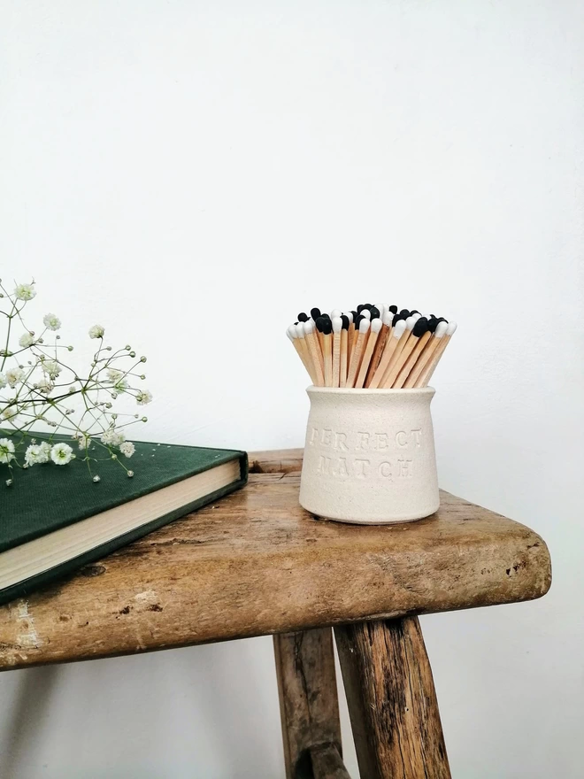 Perfect Match match pot on a wooden bench with a book and flowers. Match pot contains black and white matches