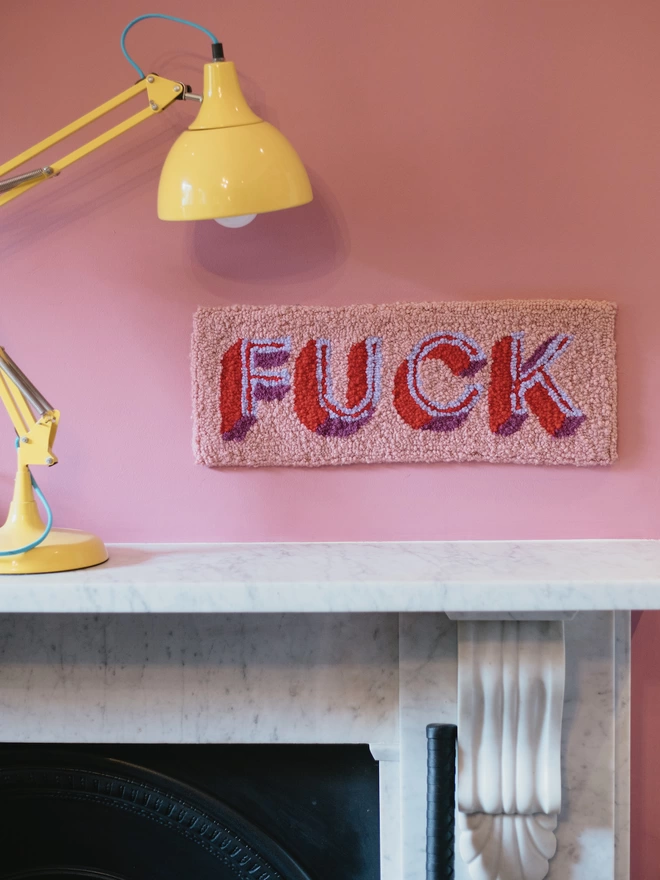 'FUCK' Handmade Tufted Rug/Wall Hanging in pink seen on a pink wall above a mantlepiece.