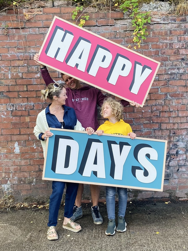 2 people and a child hold 2 wooden painted signs reading HAPPY DAYS