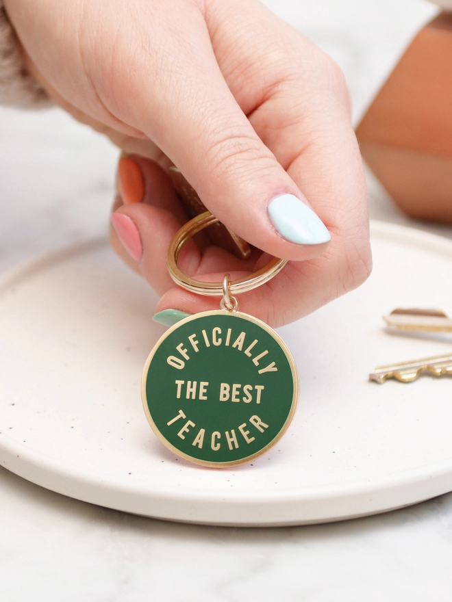 Hand holding a green keyring with 'officially the best teacher' design