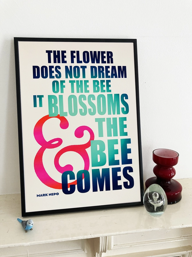 Detail from a multicoloured typographic print of “The flower does not dream of the bee, it blossoms and the bee comes” by Mark Nepo. The print sits next to a red glass candle holder, a clear glass egg and a small blue budgie.