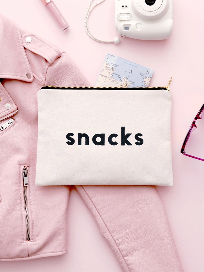 a natural canvas zip pouch with the words snacks laying on a pink background 