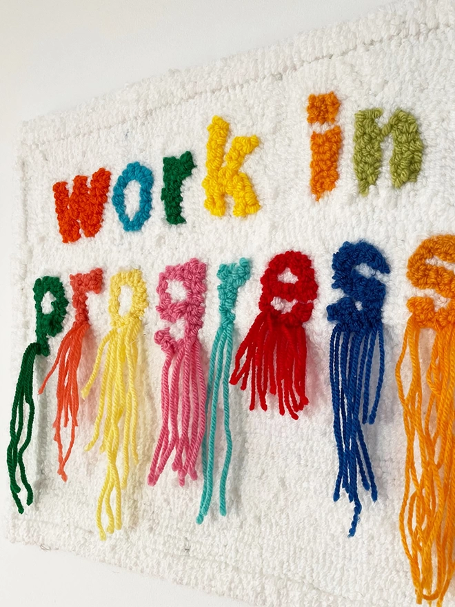 work in progress written in bright colours in wool in lower case type on a white background threads hang down from the word "progres"s