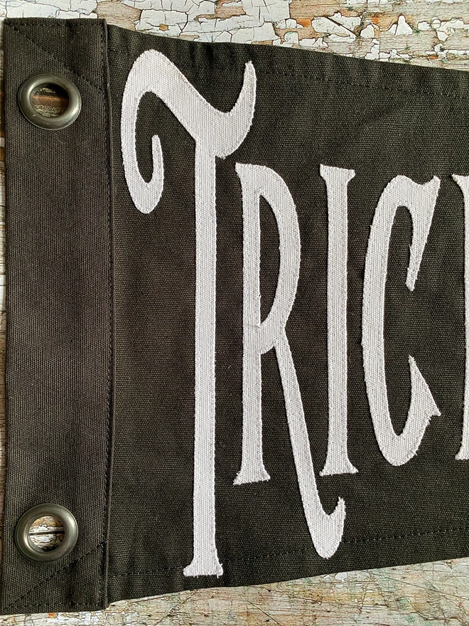 Detail of a black Trick or Tequila pennant flag. You can see the white canvas letters TRIC on black canvas.