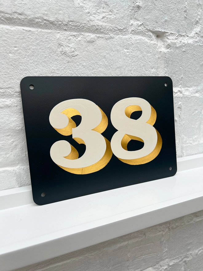 Hand painted house number against a white brick wall, number 38 in serif style in off white and directionally burnished 23 carat gold leaf, on anthracite grey background. 