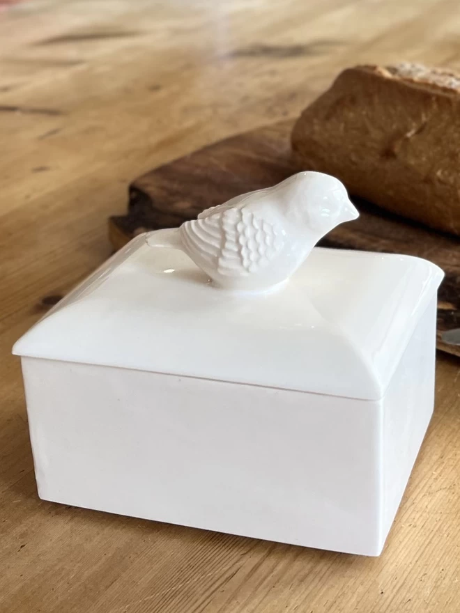A handmade ceramic butter dish lid, emulates a pat of butter in it packaging sits on an oblong base.