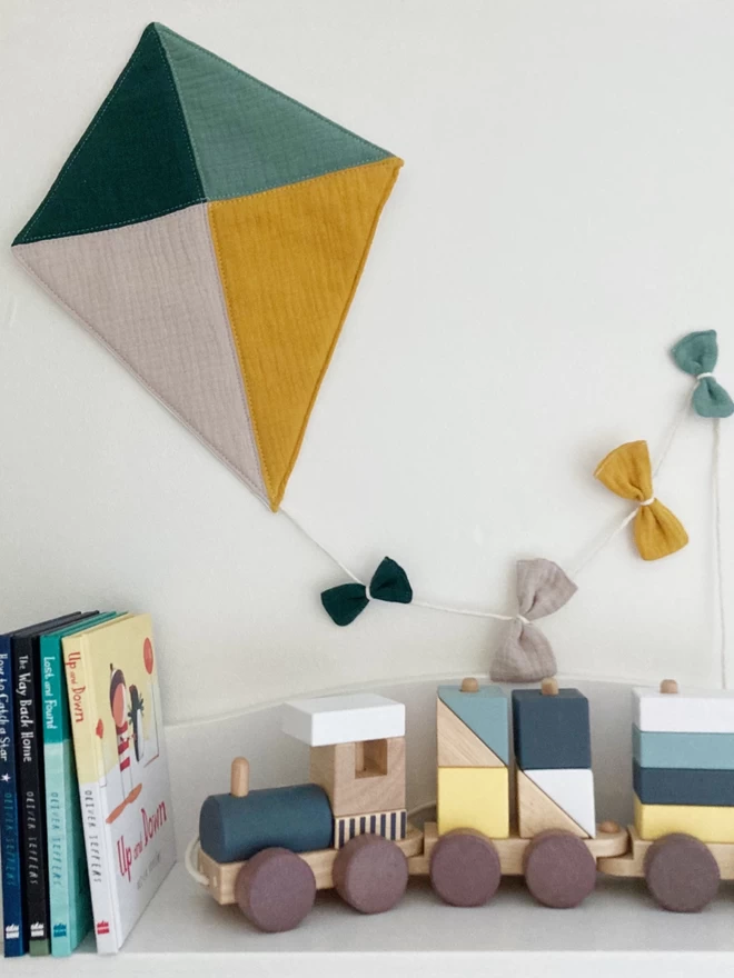 Image of the quilted kite wall hanging in green, ochre and stone by Cooper and Fred on a wall above a shelf with decorative items.