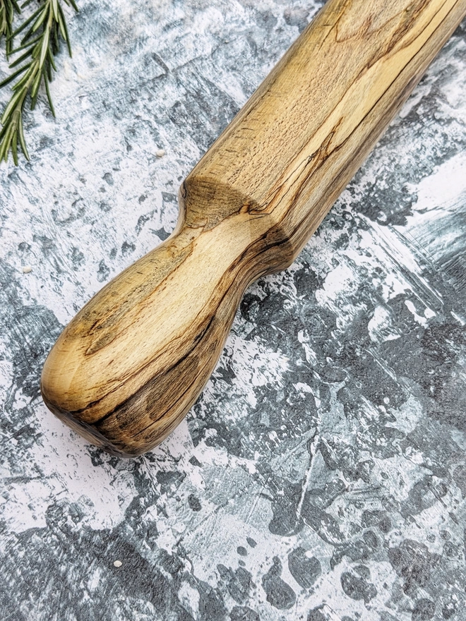 A highlight shot of a hand made rolling pin in Spalted Beech by Something From The Turnery, the picture is focussed on the handle of the rolling pin which boasts a unique darker brown grain pattern set on it’s more traditional golden Beech colour.