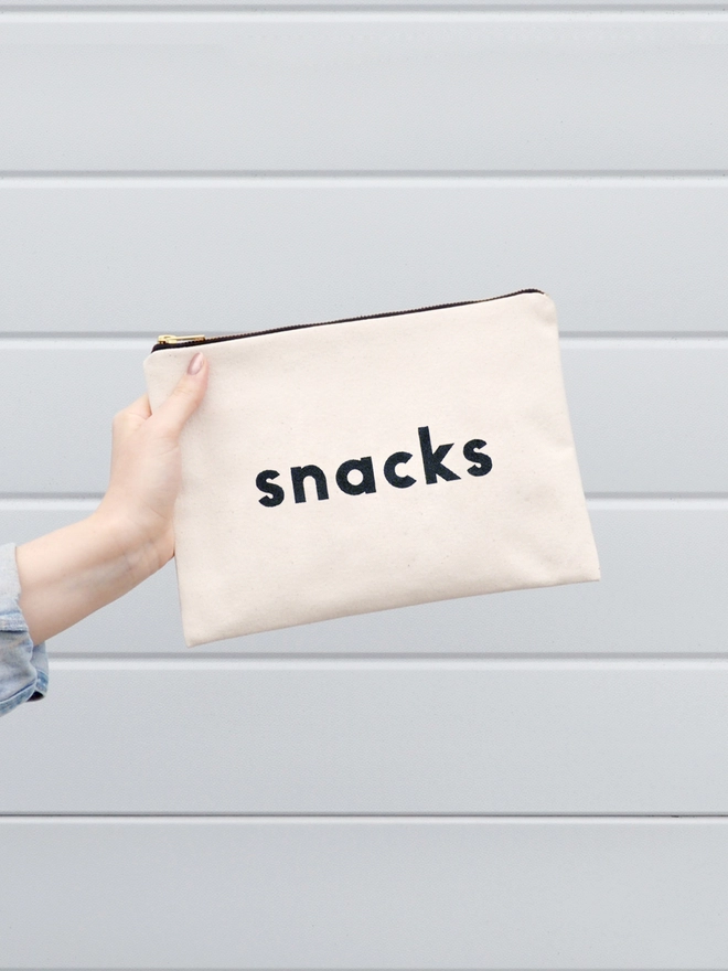 a hand holding out a natural canvas zip pouch with the words snacks