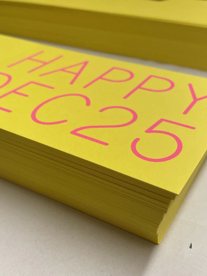 Angled view of yellow cards with neon pink wording screenprinted on top. HAPPY DEC 25 a no nonsense sentiment for Christmas greetings