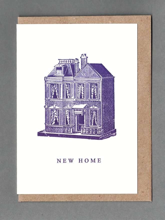 White card with purple illustration of a dolls house and text reading 'New Home' with a brown envelope behind