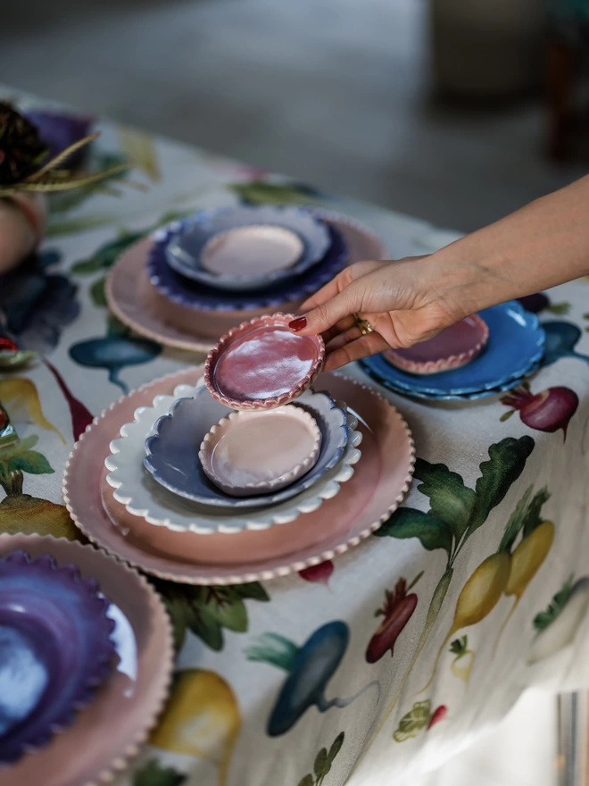 a tabletop scene with stacks of scalloped edge tableeware and a hand placing a pink coaster into the scene
