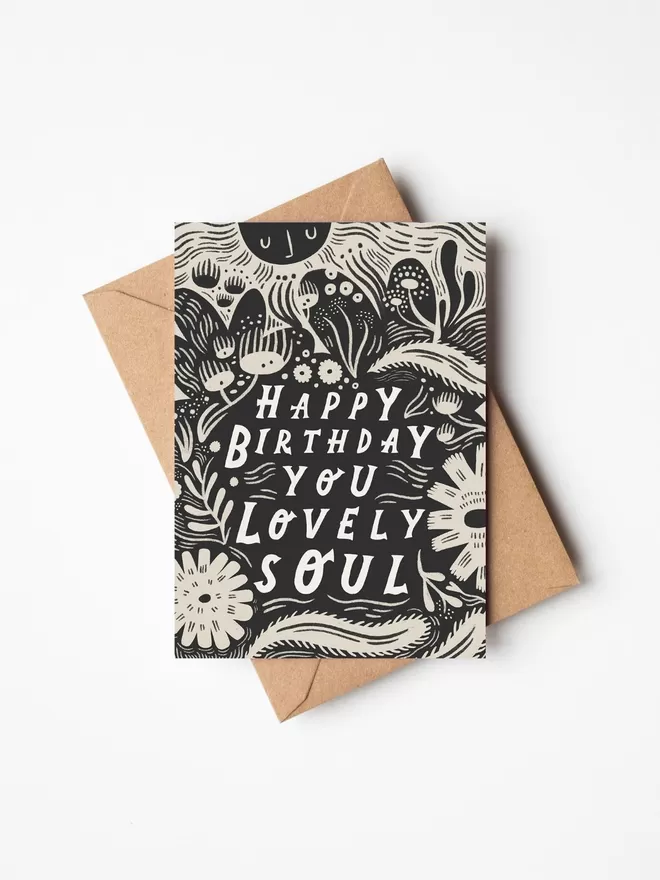 Black and white greeting card with illustration and the words Happy Birthday you lovely soul written on it with a brown envelope underneath it. 