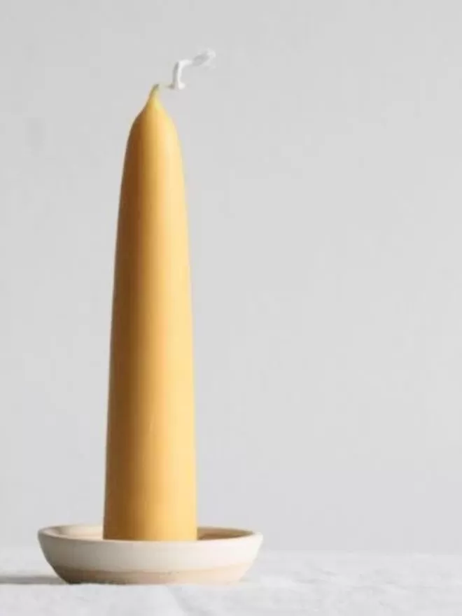 Giant stubby beeswax candle in a pearl white candle holder. Placed alone on a table top covered in white linen