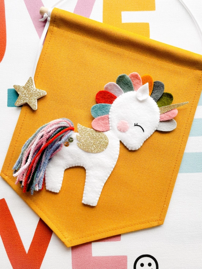 A white and gold unicorn with multicoloured mane and tail on a mustard coloured fabric banner