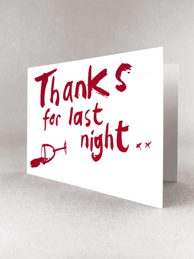 Thanks for Last night in a handwritten scrawl is screenprinted across this greetings card in claret red, with a knocked over wine glass besides. The card is stood  in a light grey studio background, slightly open.
