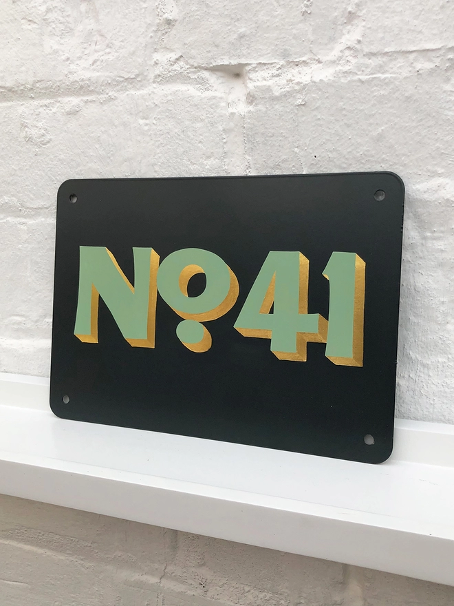 Hand painted house number No. 41 in sage green and gold leaf on anthracite grey metal plaque, against a white brick wall. 