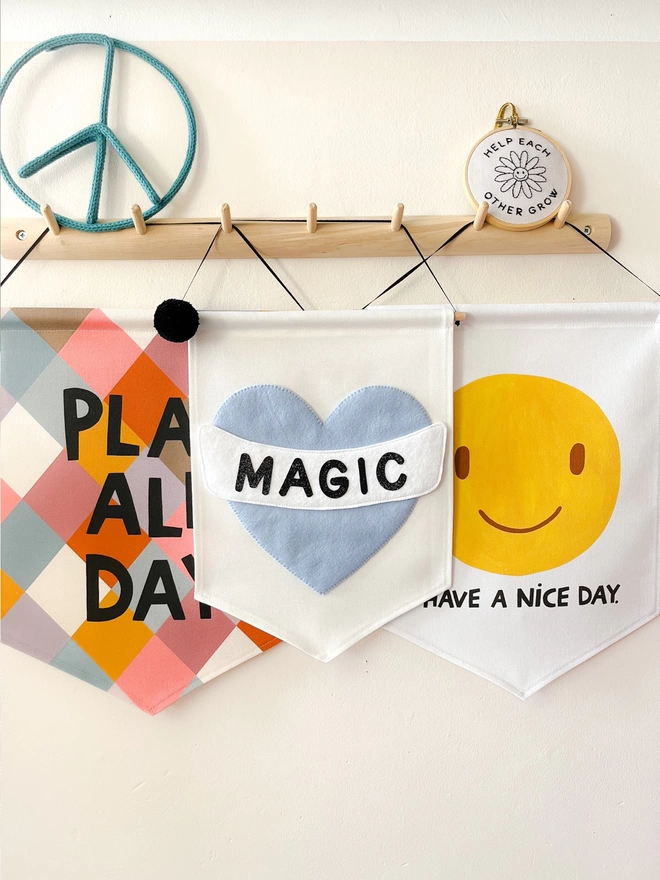 Three banners hanging from a wooden peg rail with a play all day banner magic hand sewn banner and smiley have a nice day banner