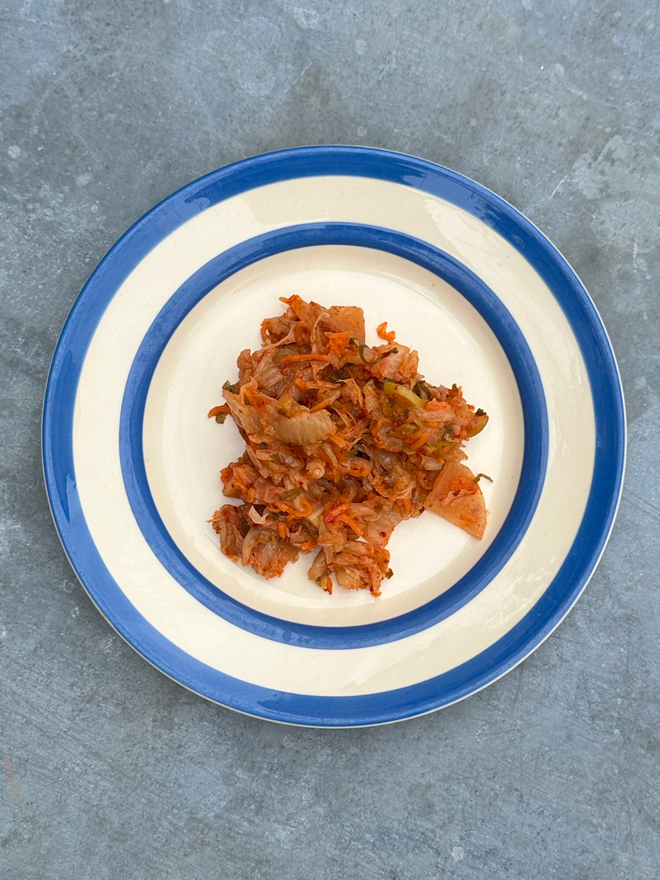 Muti Kimchi On A Blue And White Plate Atop A Steel Topped Table.