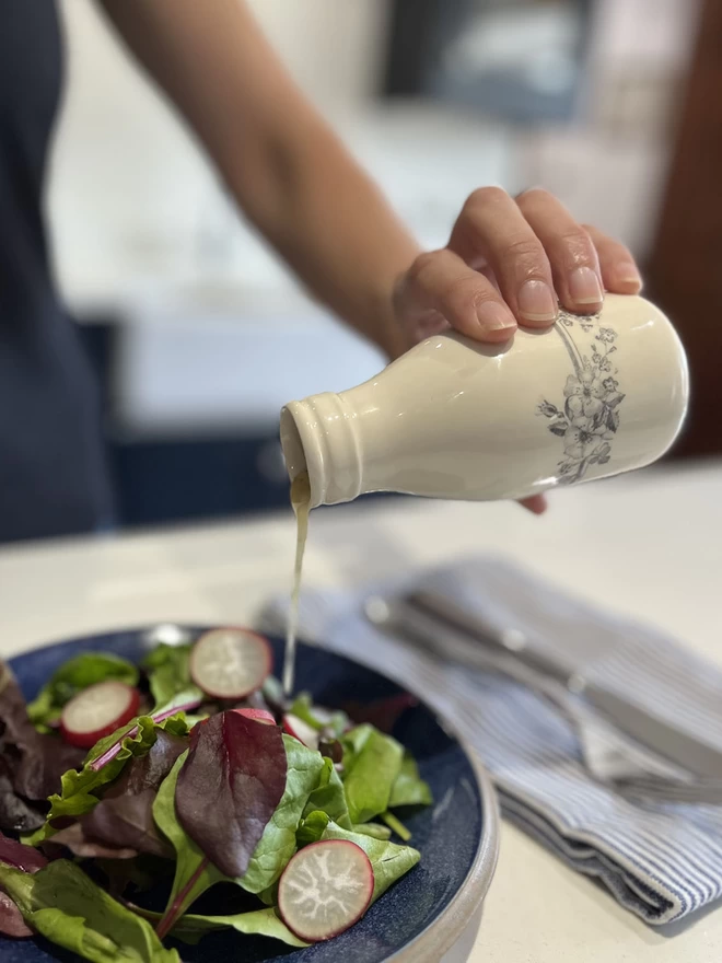  A small handmade ceramic ‘milk’ bottle/vase is pouring dressing over a plate of fresh salad. 