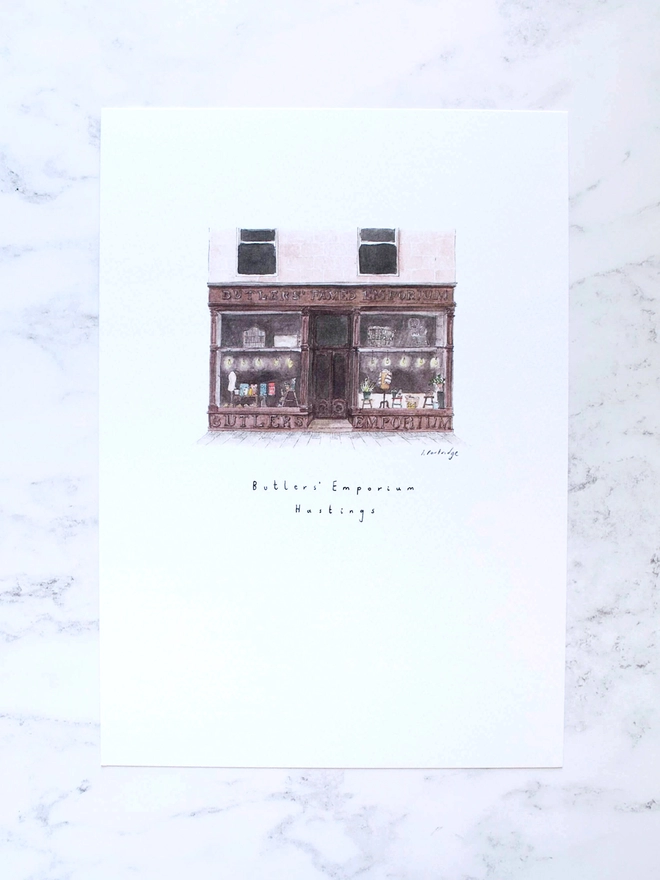 Watercolour illustration of Butler’s Emporium shop in Hastings, a brown shopfront with beautiful big windows full of treasures, the shop sells homewares and gifts carefully curated from small independent and local businesses. The painting is on a white piece of paper with the name written below. The illustration is a small watercolour painting in the centre of the white page, the page is laid out on a pale white marble background. 