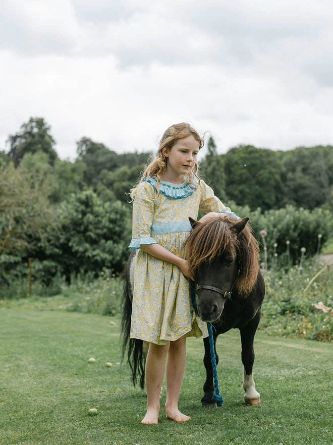 A girl stands with a miniature pony. She is wearing a yellow floral dress with a blue frill collar and sleeves and a blue sash