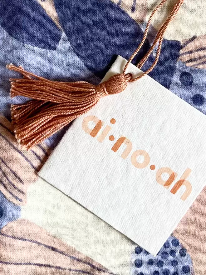 Recycled white paper tag with ainoah brand logo printed and dusty pink tassel and hanging loop