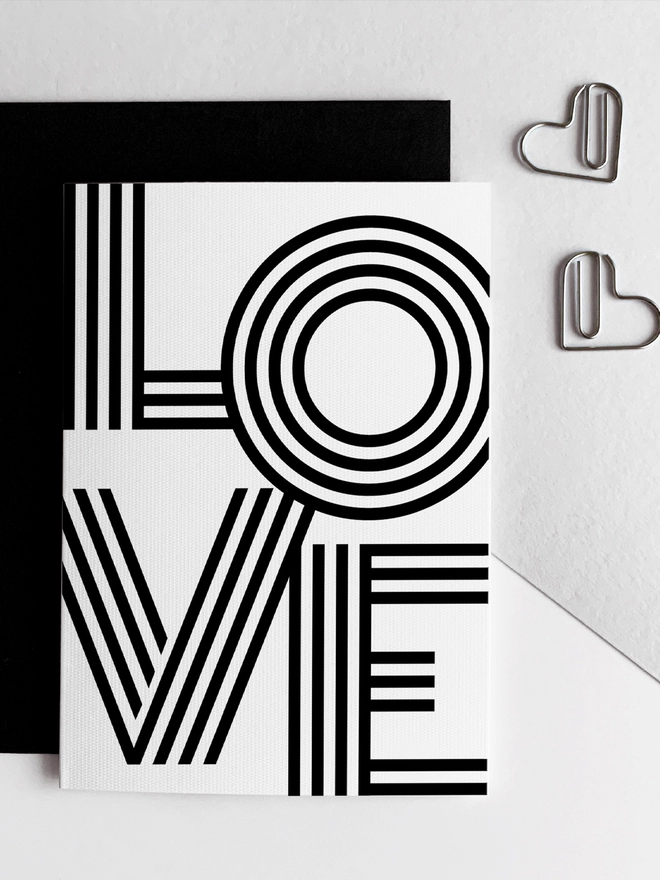 LOVE Monochrome Anniversary Card overlapping a black envelope next to heart shaped paperclips.