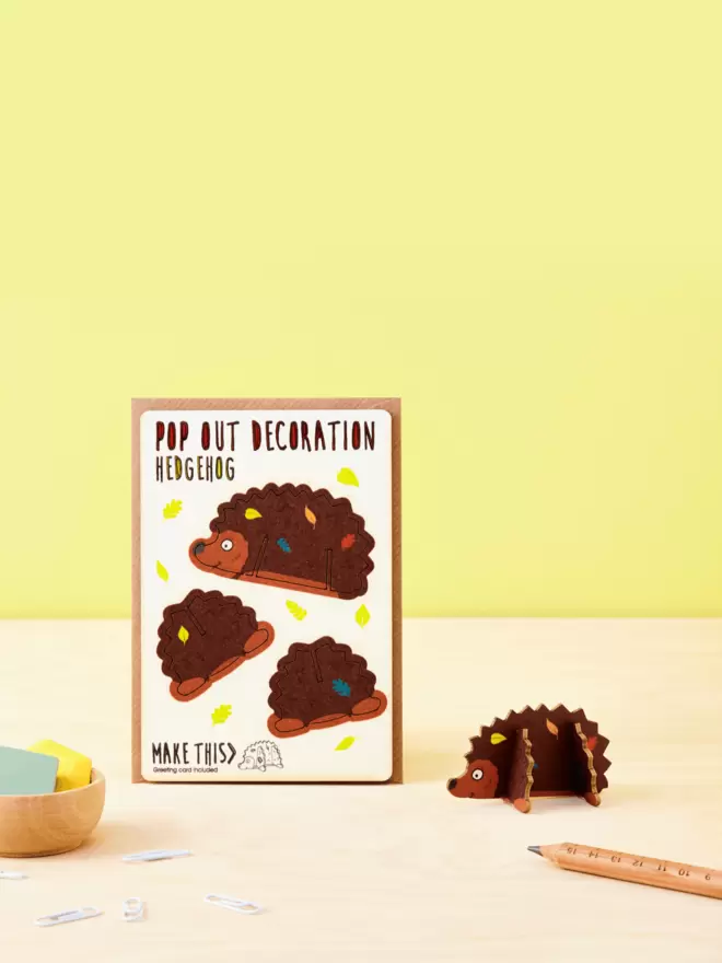 3D laser-cut hedgehog decoration and hedgehog pattern greeting card and brown kraft envelope on top of a wooden desk in front of a pale yellow coloured background