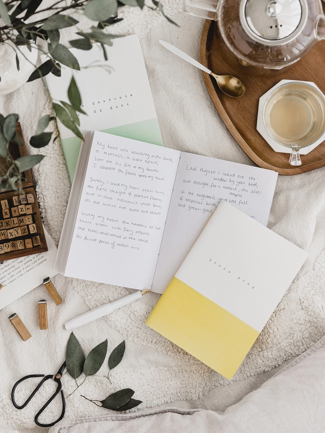 yellow and white notebook and an open notebook with writing in on a table with tea and eucalyptus
