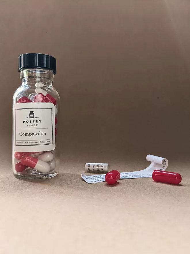 A glass bottle of red and white Compassion poetry pills, with an open pill displaying the rolled quote it contained