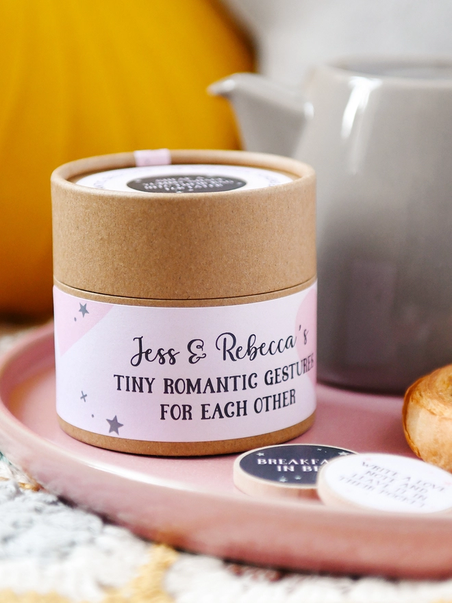 A cardboard jar with a pale pink label that reads 'Tiny romantic gestures for each other' stands on a pink plate beside a teapot and two wooden tokens.