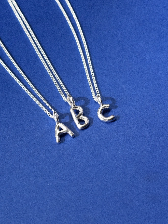three silver alphabet pendants, A, B, C, on thin silver chains laid on a royal blue background.