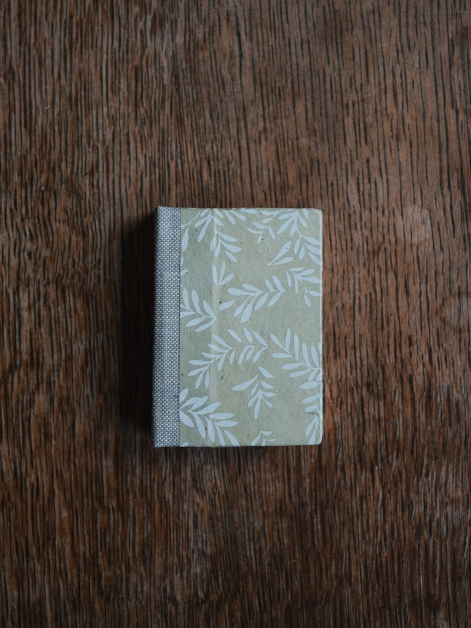 Miniature book with a pale green cover with white leaf pattern and beige cloth spine