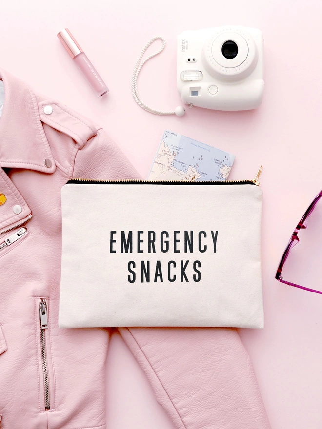 a natural canvas zip pouch with the words emergency snacks laying on a pink backdrop with travel items surrounding