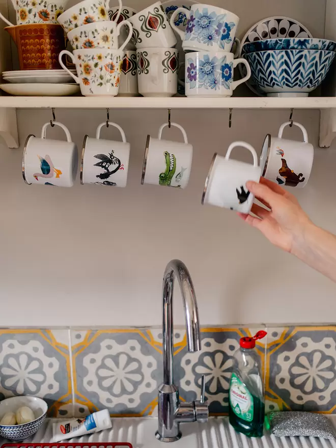 A hand reaches to take a decorated enamel mug off a hook over a sink