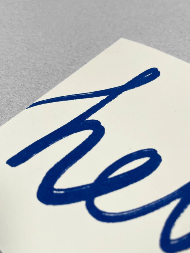 Close up of a handwritten blue hello, screenprinted on this landscape off-white greetings card. 