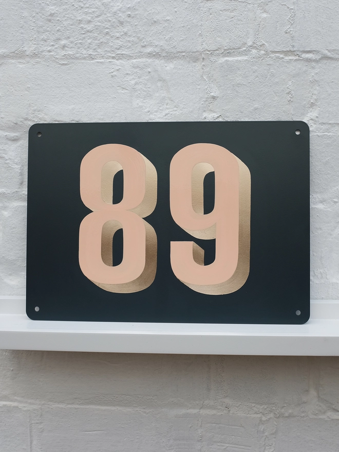 Peach and gold leaf house number 89, on anthracite grey metal plaque, against a white brick wall.