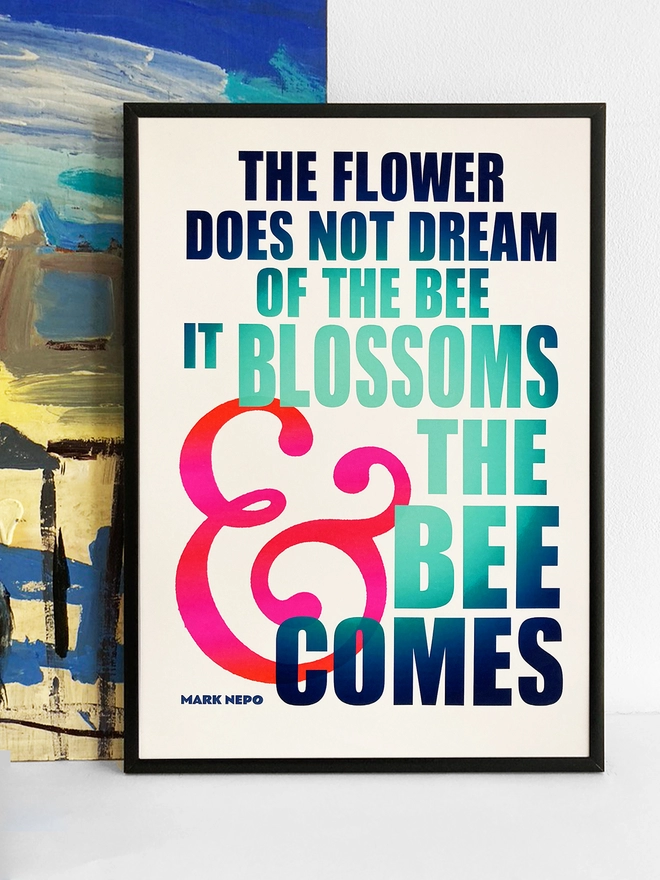 Framed multicoloured typographic print of “The flower does not dream of the bee, it blossoms and the bee comes” by Mark Nepo.  The print rests against a blue and yellow abstract painting.
