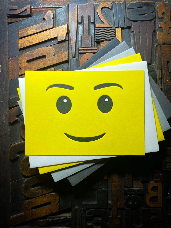 A bright yellow letterpress card with a smiling face emoji is placed on top of a selection of contrasting envelopes. The background consists of various wooden printing letters in different orientations and sizes.